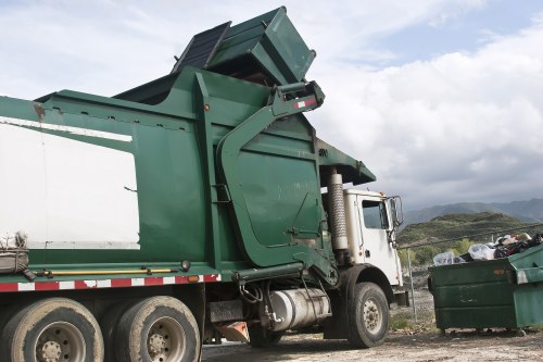 A large industrial waste collection truck with a green metal body and a white cab, parked outdoors on a gravel surface. The truck's rear compaction mechanism is raised, revealing an open hopper for loading rubbish. The side panels are made of textured metal with visible seams and rivets, painted in green with some signs of weathering. To the right of the truck, there is a dark green plastic skip filled with mixed waste items, including black bin bags and assorted debris, placed on the ground beside the gravel. The background features a partly cloudy sky with patches of blue and distant hilly terrain, suggesting a rural or semi-industrial area. The scene indicates a rubbish collection or waste removal operation, typical of services offered by companies such as Business Waste Removal Knightsbridge, servicing the London postcode area including Knightsbridge.