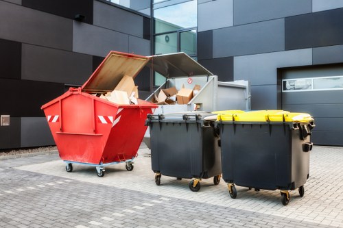 In the image, there are three large wheeled rubbish bins positioned on a paved outdoor area. The first bin, on the left, is a red skip with an open top, revealing a variety of cardboard boxes and packaging materials inside, with some boxes leaning against the sides. The surface of the red bin appears metallic with a smooth finish and red-and-white reflective strips on the back. Adjacent to it, slightly to the right, are two black wheelie bins with yellow lids, also made of plastic, standing side by side. The black bins have a textured surface typical of plastic waste containers and are fitted with sturdy wheels at their base for mobility. The background features a modern building with dark, rectangular panels and large glass windows, indicating an urban or commercial setting, possibly in Knightsbridge. The scene is well-lit with natural daylight, and the environment suggests an area designated for waste management or rubbish collection, consistent with services offered by Business Waste Removal Knightsbridge.