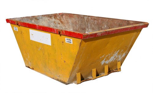A large industrial skip situated outdoors on a paved surface, primarily yellow in colour with a red rim around its top edge, which shows rust and dirt accumulation. The skip's sides are slightly tapered and display signs of wear, including scratches and chipped paint, indicating frequent use. The interior of the skip is visible and appears to be made of a rough, metallic material with a layer of dirt and debris. The skip is positioned in an open environment with no other objects or vehicles nearby, and faint natural light illuminates the scene. This type of skip is commonly used for commercial waste removal, including bulky refuse and building debris, aligning with services provided by Business Waste Removal Knightsbridge. The skip's sturdy construction and heavy-duty design are suitable for waste collection in urban areas, and its placement suggests it is ready for loading or recent use in a waste management process in London postcodes, such as SW1.