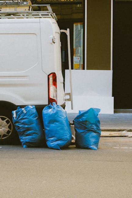 The image shows the rear side of a white commercial van parked on a street, with three large, blue, plastic rubbish bags placed on the pavement directly behind it. The bags appear to be filled with wa