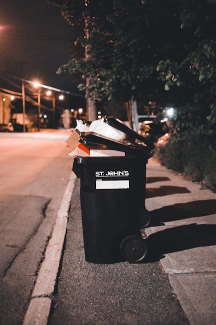 A row of five large, white plastic waste bins with closed hinged lids are positioned at the edge of a paved outdoor area, likely a driveway or service yard, in front of a dark-colored shipping contain